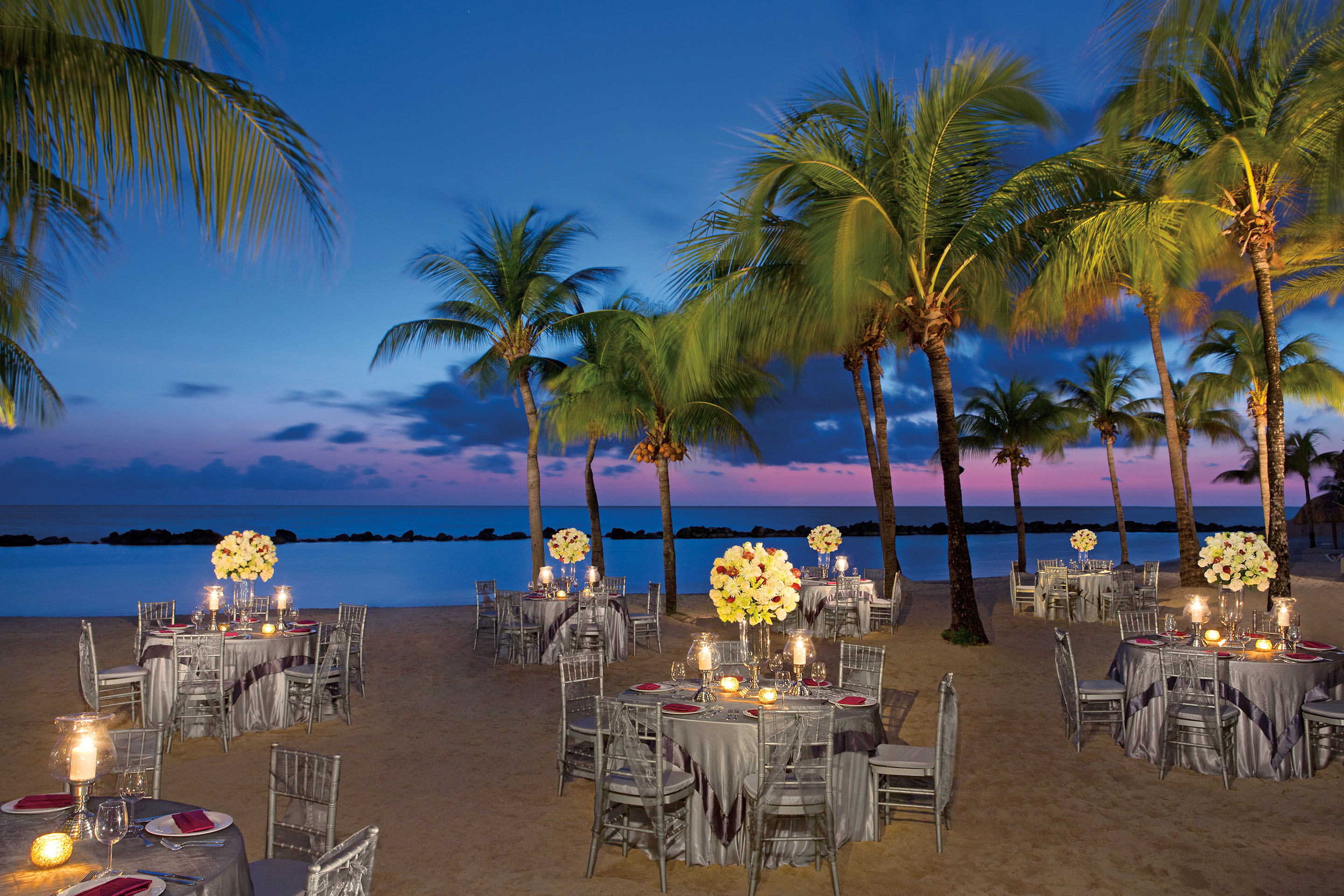 Caribbean beach set with elegant sit-down tables surround by palm trees in early evening